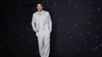 La imagen muestra a un hombre vestido con un traje claro de color gris. El traje tiene un corte elegante, con pantalones de tiro alto y una camisa debajo que parece ser de un tono similar. El hombre tiene una expresión sonriente y está posando de manera confiada, con una mano en el bolsillo del pantalón. El fondo es oscuro y está adornado con luces brillantes que aportan un toque festivo a la escena.