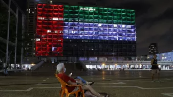 En la imagen se puede ver a un hombre mayor sentado en una silla de plástico anaranjada, mirando su teléfono móvil. Detrás de él, se destaca un gran edificio iluminado que muestra los colores de la bandera de México: rojo, blanco y verde. Es de noche, y el ambiente parece tranquilo con algunas personas caminando en el área. La arquitectura del edificio se aprecia moderna, y las luces crean un contraste vibrante con el cielo nocturno.