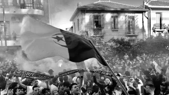 La imagen muestra a una gran multitud en un ambiente festivo, probablemente durante un evento deportivo o una celebración. Se pueden ver personas ondeando una bandera, así como pancartas y bufandas, lo que sugiere un fuerte sentido de identidad y apoyo a un equipo. El ambiente está lleno de humo, lo que añade un efecto dramático a la escena. La gente parece emocionada y unida, disfrutando del momento. Las edificaciones al fondo indican que la actividad está ocurriendo en un área urbana.