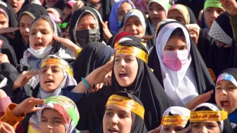 La imagen muestra un grupo de niñas y jóvenes con pañuelos en la cabeza y cintas en la frente. Están reunidas en un evento, posiblemente una manifestación o celebración, y parecen estar cantando o gritando algo en un ambiente lleno de energía. Las niñas lucen diversas expresiones de emoción y entusiasmo, mientras que algunas llevan mascarillas. La diversidad de colores en sus ropas y cintas resalta un sentido de unidad y propósito en su actividad.