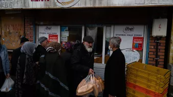 La imagen muestra a un grupo de personas en la calle, frente a una tienda. Algunos de ellos llevan bolsas en las manos, probablemente con pan. La escena sugiere una atmósfera de actividad comunitaria, ya que la gente está haciendo fila. Se pueden ver diferentes edades y estilos de ropa entre los presentes. Además, hay un letrero visible en la tienda que indica que se trata de un punto de venta de pan en Estambul.