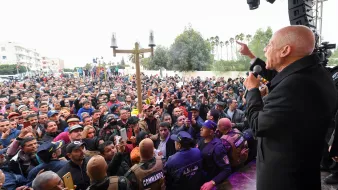 En la imagen se observa a un grupo numeroso de personas reunidas, probablemente en un evento o mitin. En el frente, hay un orador que está hablando con entusiasmo, utilizando un micrófono. Al fondo, se pueden ver elementos de seguridad, como un grupo de personas con chalecos que indican su función de protección o control del evento. El ambiente parece ser de gran expectación, y el público muestra interés en lo que se está diciendo. Además, hay un escenario o tarima donde se encuentra el orador. La escena transmite una sensación de participación activa de la gente.