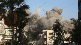 La imagen muestra una escena de destrucción y caos en una ciudad. Se puede ver una gran nube de polvo y escombros elevándose en el aire, lo que sugiere que ha ocurrido una explosión o un colapso de un edificio. En primer plano, hay un grupo de personas que parecen estar observando la situación, algunas de ellas con expresiones de sorpresa o preocupación. Alrededor, hay palmeras y estructuras urbanas que indican que se trata de un área urbana. El cielo es de un color azul claro, contrastando con el humo gris.