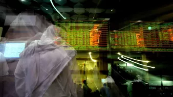 La imagen muestra un ambiente vibrante de una bolsa de valores o un mercado financiero. Se pueden ver pantallas con información en tiempo real, incluyendo gráficos y precios en verde y rojo, que indican cambios en el mercado. En primer plano, hay figuras en movimiento que parecen ser personas vestidas con ropa tradicional, lo que sugiere un contexto cultural específico. La imagen transmite una sensación de dinamismo y actividad intensa, típica de un entorno bursátil.
