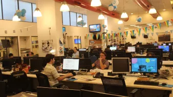 La imagen muestra una oficina moderna y espaciosa. Hay varias estaciones de trabajo con computadoras y pantallas en escritorios. Algunas personas están concentradas en sus tareas mientras que otras parecen colaborar o comunicarse. El ambiente es iluminado y decorado con globos y banderas, lo que sugiere que podría ser un lugar festivo o creativo. Las paredes tienen fotografías y otros elementos decorativos, añadiendo un toque personal al espacio laboral.