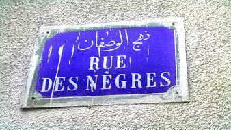 La imagen muestra una placa de calle con un fondo azul oscuro. En la placa se encuentran tres inscripciones en diferentes idiomas: árabe, francés y otro que parece ser un nombre transliterado. La parte central dice "RUE DES NÈGRES" en letras blancas. La placa parece estar un poco desgastada y sucia, lo que indica que ha estado expuesta al tiempo.