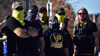 La imagen muestra a un grupo de hombres con vestimenta oscura, algunos de ellos llevan pañuelos o mascarillas en la cara. Están de pie, en una actitud seria, y parecen estar en una manifestación o evento público. Hay una mezcla de gafas de sol y atuendos que sugieren un sentido de unidad o pertenencia a un grupo específico. El ambiente general indica que pueden estar participando en una actividad de interés político o social.