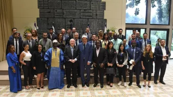 La imagen muestra a un grupo de personas posando juntas en un espacio interior bien iluminado. Al fondo, se pueden ver banderas y una pared decorativa. El grupo incluye hombres y mujeres de diversas apariencias, algunos con trajes formales y otros con atuendos tradicionales. Todos están sonriendo y parecen reunidos para una ocasión especial, posiblemente un evento diplomático o cultural. La disposición del grupo sugiere una atmósfera de cooperación y celebración.