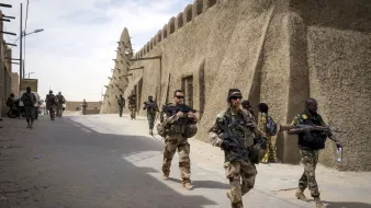 La imagen muestra a un grupo de soldados que caminan por una calle en una zona con arquitectura de barro. A su alrededor, se pueden ver estructuras típicas del lugar, como muros de adobe. Los soldados llevan equipos militares y armamento, y algunos parecen estar en formación o realizando patrullas. La escena sugiere un ambiente de seguridad o una operación militar en un contexto urbano o semiurbano.