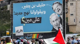 La imagen muestra una gran pancarta en un evento, donde destacan las imágenes de dos personas, una de ellas es un hombre que parece ser una figura política conocida. El cartel contiene texto en árabe, con un mensaje crítico, probablemente relacionado con la situación política. En primer plano, se observan personas reunidas, algunas de las cuales sostienen banderas, sugiriendo que puede ser una manifestación o protesta. La atmósfera parece ser de movilización social.