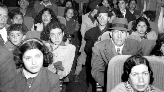 La imagen muestra el interior de un avión lleno de pasajeros. Hay muchos hombres, mujeres y niños sentados en los asientos con expresiones variadas. La mayoría de las personas parecen estar mirando hacia la cámara. El ambiente es de una época pasada, con un estilo de vestimenta que refleja un tiempo diferente. La iluminación es tenue, lo que contribuye a la atmósfera general de la fotografía.