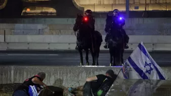 La imagen muestra a un par de oficiales montados sobre caballos, con luces rojas y azules, vigilando una situación que parece estar relacionada con una protesta. En el primer plano, hay dos personas en el suelo, una de las cuales sostiene una bandera de Israel. La escena parece indicar una tensión o un conflicto en el contexto de la protesta, con un ambiente urbano de fondo, probablemente en una carretera. La iluminación y la posición de los agentes reflejan la seriedad de la situación.