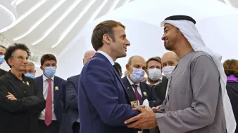 En la imagen se pueden ver a varias personas en un ambiente formal. Al frente, dos hombres están estrechando manos, uno de ellos viste un traje y el otro lleva una vestimenta tradicional árabe. Alrededor de ellos, hay más personas que observan la interacción, algunas de las cuales llevan mascarillas. El fondo muestra una arquitectura moderna y luminosa. La atmósfera es de cordialidad y diplomacia.