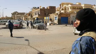 La imagen muestra una escena urbana donde varios agentes de seguridad están presentes en una calle. Algunos de ellos están uniformados y parecen estar patrullando la zona. Hay vehículos estacionados, incluidos autos civiles y un todoterreno. El ambiente parece tenso, y hay edificaciones a los lados de la calle que sugieren un entorno urbano. La luz del día indica que es probablemente por la mañana o la tarde.