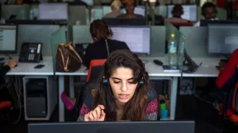La imagen muestra un ambiente de oficina, específicamente un centro de llamadas. En primer plano, hay una mujer con un auricular que parece estar concentrada en su trabajo, hablando y escribiendo en su computadora. Detrás de ella, se pueden ver varias personas trabajando en sus estaciones, también usando auriculares y frente a pantallas. La iluminación es brillante y moderna, y cada escritorio tiene un teléfono y botellas de agua. La escena transmite una sensación de trabajo dinámico y colaborativo.