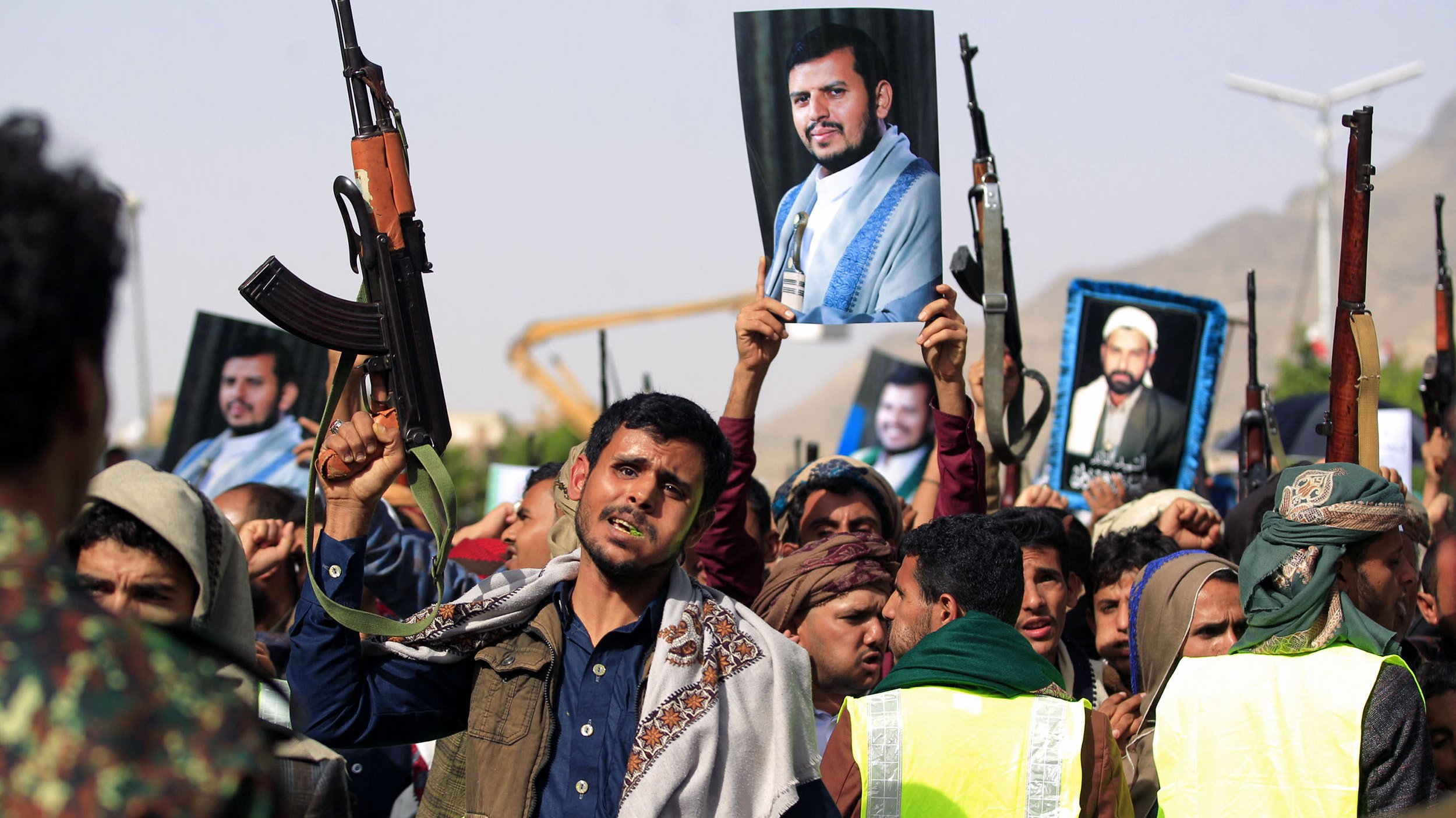 در این تصویر، گروهی از مردم در حال برگزاری یک تجمع هستند. تعدادی از شرکتکنندگان سلاحهای خود را بلند کردهاند و تعدادی دیگر تصاویری از یک فرد را در دست دارند. این تجمع به نظر میرسد که از جنبه سیاسی و اجتماعی برخوردار باشد. مردمی که در تصویر دیده میشوند، لباسهای سنتی و محلی به تن دارند و برخی از آنها روسریهایی به سر بستهاند. پسزمینه تصویر شامل تعدادی سازه و فضای باز است.