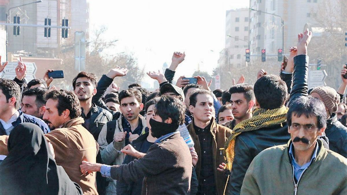 در این تصویر، گروهی از مردم در یک جمعیت انبوه دیده میشوند که به نظر میرسد در حال برگزاری یک تجمع یا اعتراض هستند. برخی از افراد دستهای خود را بالا بردهاند و دیگران به سمت دوربین نگاه میکنند. تعدادی از افراد لباسهای معمولی بر تن دارند و در پسزمینه، ساختمانها و چراغهای راهنمایی قابل مشاهده است. حالت چهرهها نشاندهندهی هیجان یا تمایل به ابراز نظر است. برخی از افراد نیز با پوشش صورت دیده میشوند.