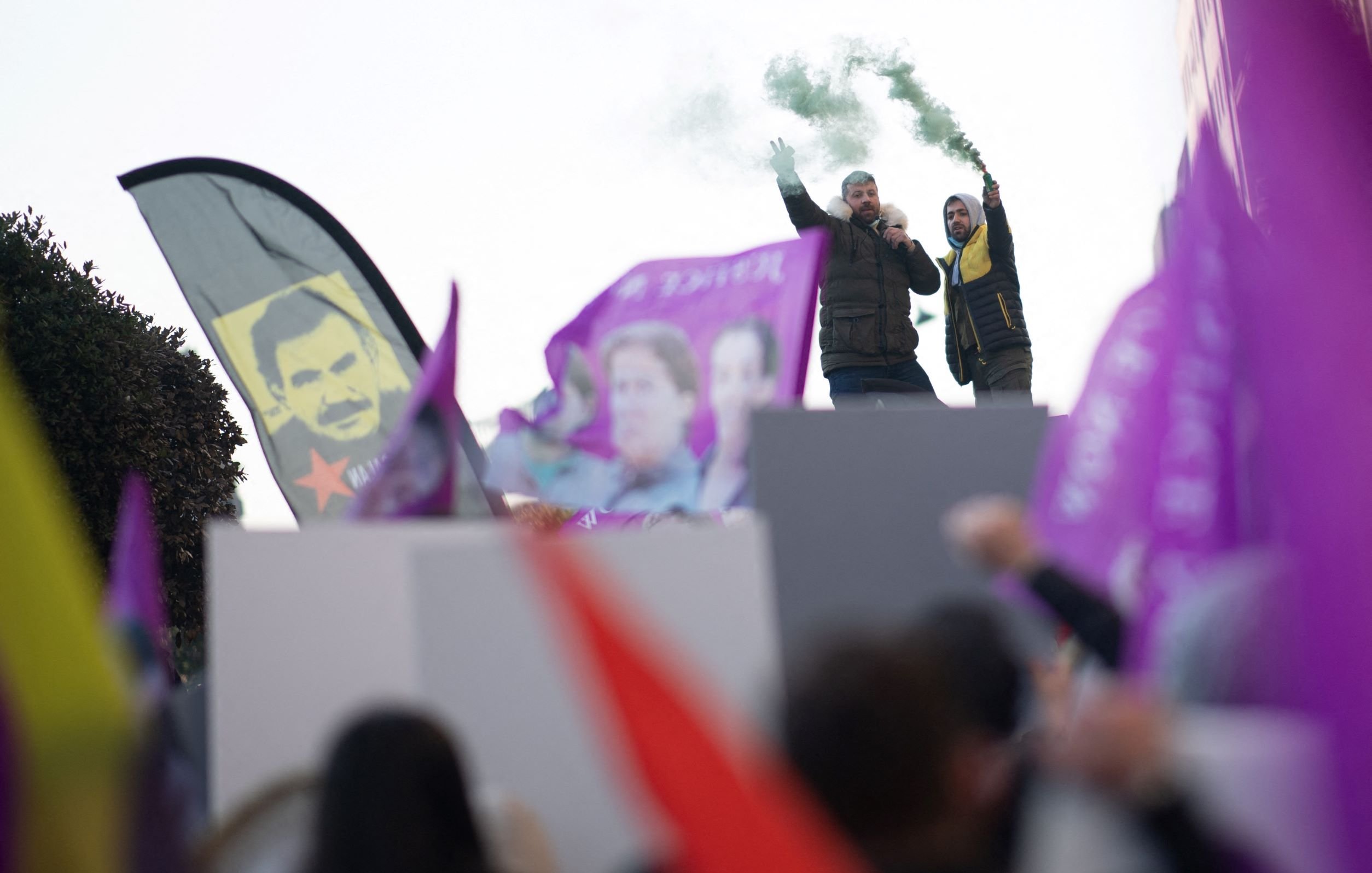 در این تصویر، تعدادی از افراد در حال برگزاری یک تجمع اعتراضی یا حمایتی به نظر میرسند. دو نفر بر روی یک سکو ایستادهاند و یکی از آنها میکروفون در دست دارد و به جمعیت صحبت میکند. در دست برخی از افراد پرچمها و بنرهایی با رنگهای مختلف، از جمله بنرهای بنفش، در حال تکان خوردن است. همچنین، دود سبز رنگی به هوا بلند شده که به جو تجمع حال و هوای خاصی میبخشد. در پسزمینه، تابلوی چهرهای نیز قابل مشاهده است.