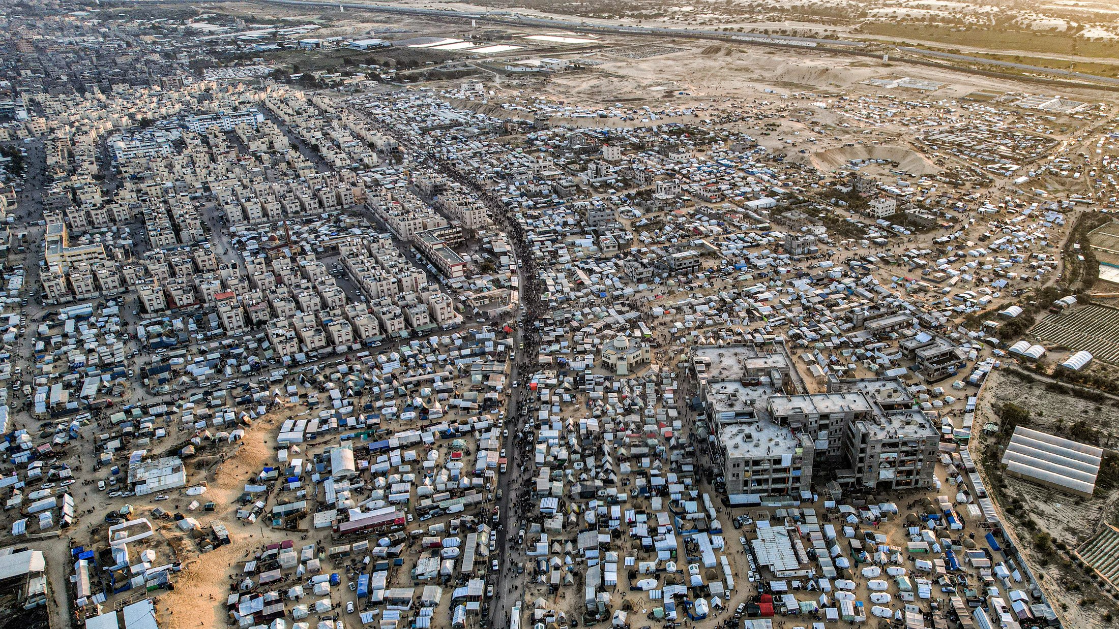 این تصویر نمای هوایی از یک منطقه شلوغ و پرجمعیت است. در اینجا تعداد زیادی ساختمان و خانه های کوچک دیده میشود که در کنار هم قرار گرفتهاند. خیابانهایی که به نظر میرسد مردم در آن تردد دارند، میانه تصویر را تقسیم کردهاند. همچنین، در برخی نواحی، چادرها و سازههای موقت دیده میشوند که نشاندهنده زندگی در شرایط سخت و غیرفرهنگین است. این تصویر، وضعیتی را به نمایش میگذارد که بیانگر چالشهای اجتماعی و اقتصادی است.