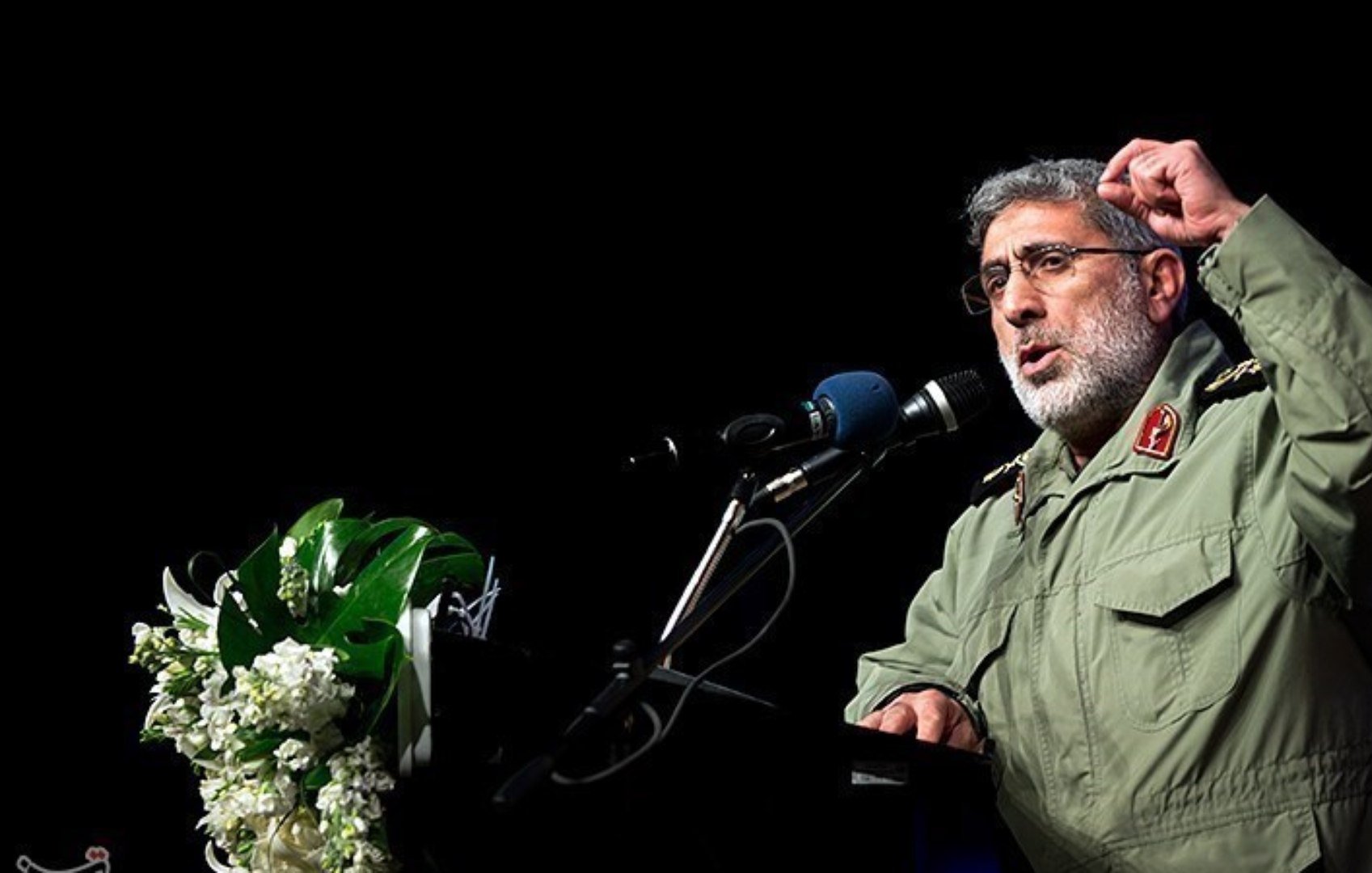 در این تصویر، یک فرد در حال سخنرانی در یک مکان رسمی است. او لباس نظامی به تن دارد و در حال بیان نکاتی با حرکات دست است. در کنار او، میزی با گلهای تزئینی قرار دارد و میکروفون و بلندگو نیز دیده میشود. پسزمینه تصویر تاریک است و تمرکز بیشتری بر روی سخنران وجود دارد.