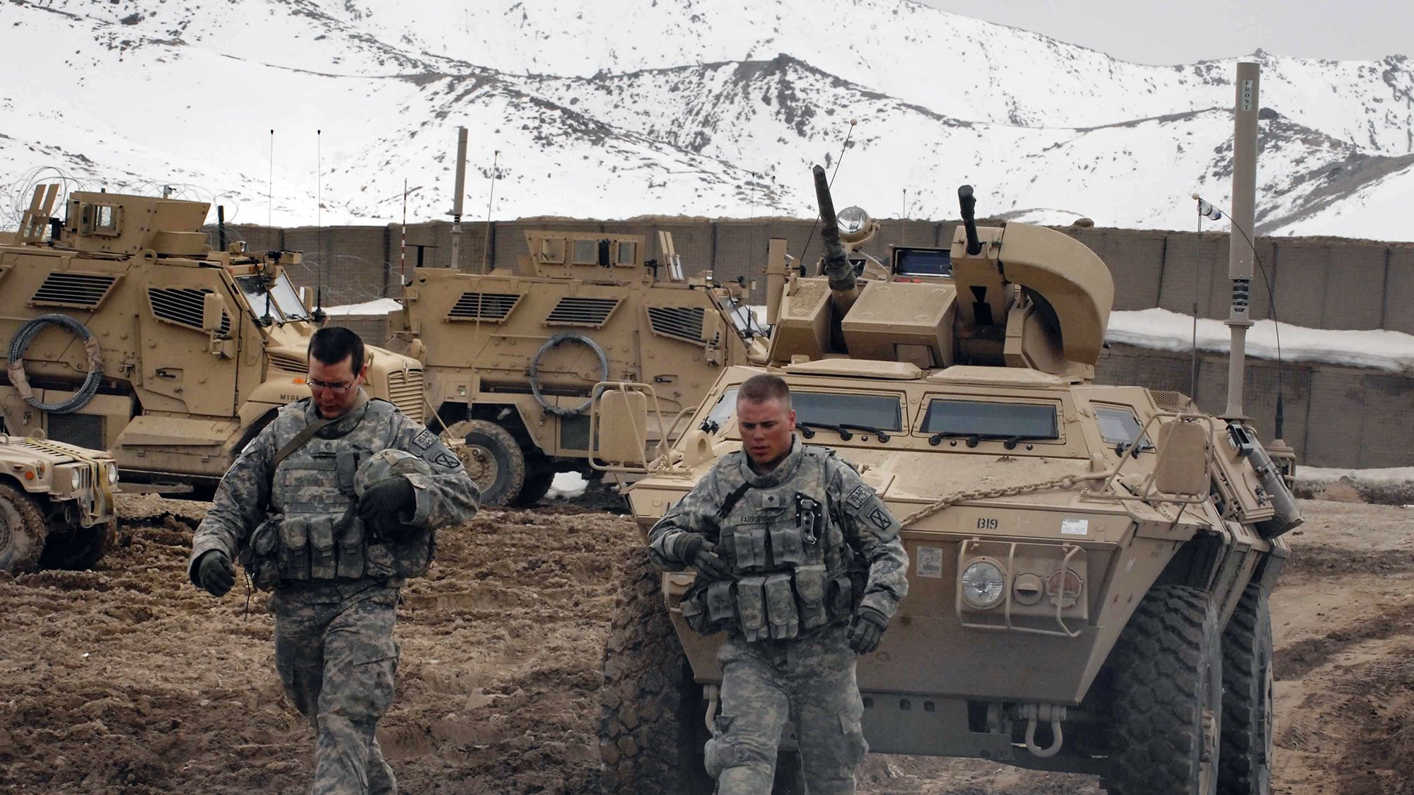 در این تصویر، دو سرباز در حال حرکت در یک زمین خاکی و سرد هستند. آنها لباس نظامی به تن دارند و در پسزمینه، چندین خودروی زرهی مشاهده میشود. کوهها و برف در دوردست قابل مشاهده است که نشاندهنده شرایط جغرافیایی سخت منطقه است. سربازان به نظر میرسد که در حال انجام یک ماموریت یا گشتزنی هستند.