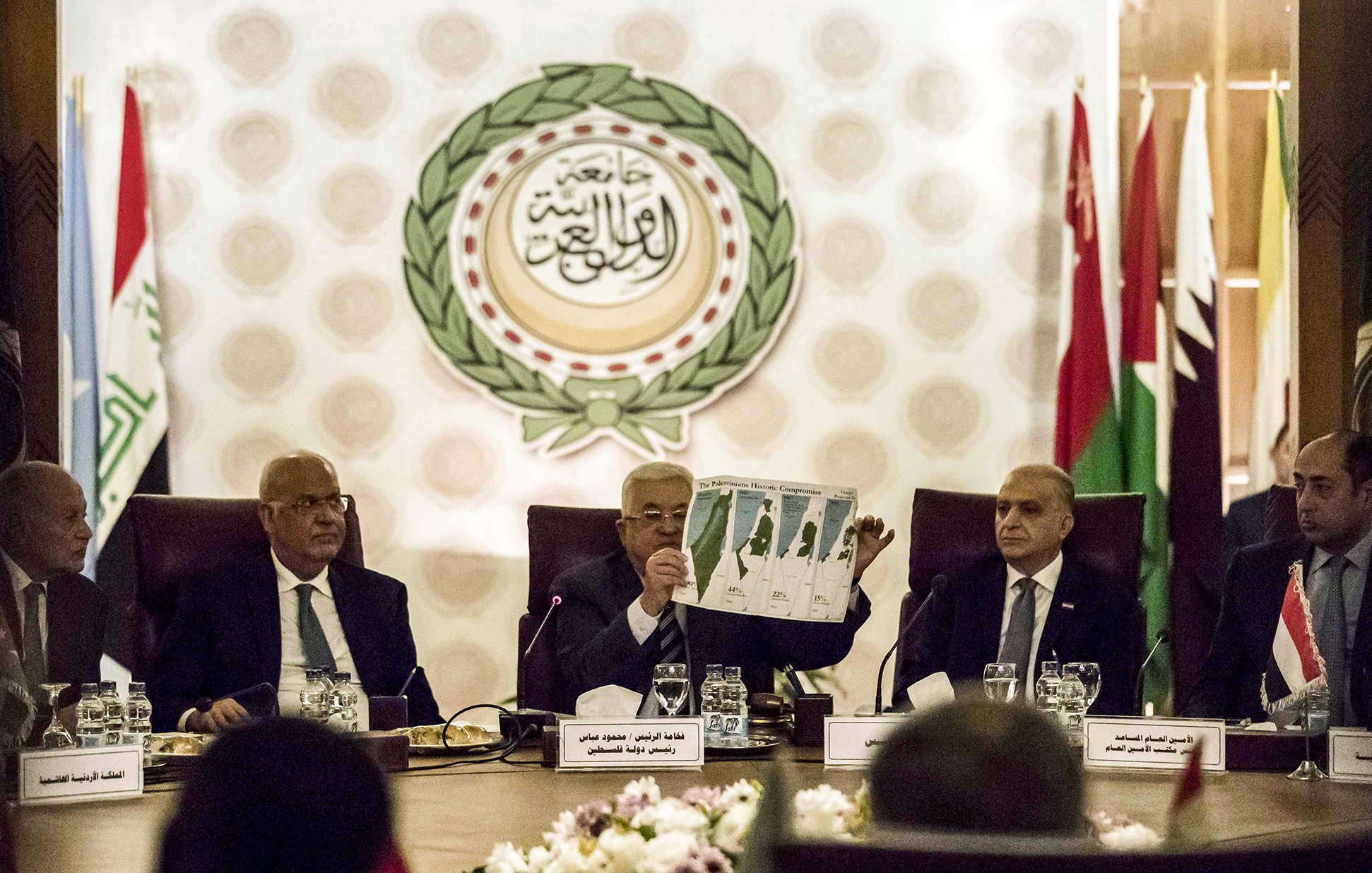 در این تصویر، گروهی از افراد در یک جلسه رسمی نشستهاند. یکی از افراد در حال نشان دادن یک نقشه یا نمودار به دیگران است. پشت آنها پرچمهای کشورهای مختلف و نماد یا لوگوی اتحادیه عرب قابل مشاهده است. فضای جلسه حرفهای و سازمانیافته به نظر میرسد. اعضای حاضر به دقت به موضوع مورد بحث گوش میدهند.