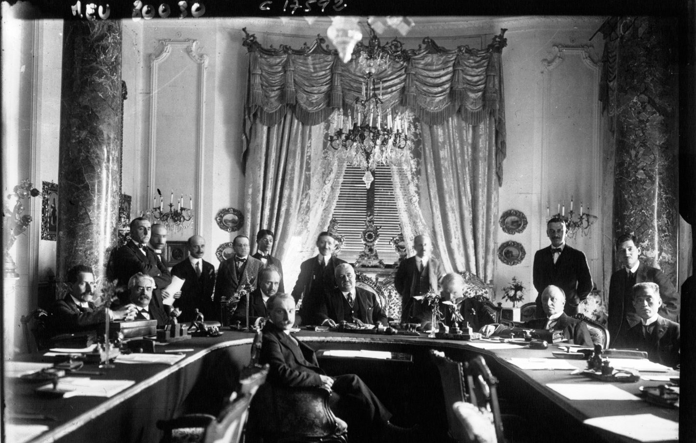 این تصویر، یک جلسه یا کنفرانس تاریخی را نشان میدهد که در آن گروهی از مردان در یک اتاق بزرگ و با طراحی تجملاتی گرد هم آمدهاند. در مرکز تصویر، یک مردی با لباس رسمی نشسته است و دیگر افراد دور او ایستاده و نشستهاند. دکوراسیون اتاق شامل پردههای بلند، لوستر و تزئینات دیواری است که به فضای جلسه شکوه و جلال خاصی میبخشد. این تصویر نشاندهنده یک رویداد مهم و رسمی در تاریخ است.