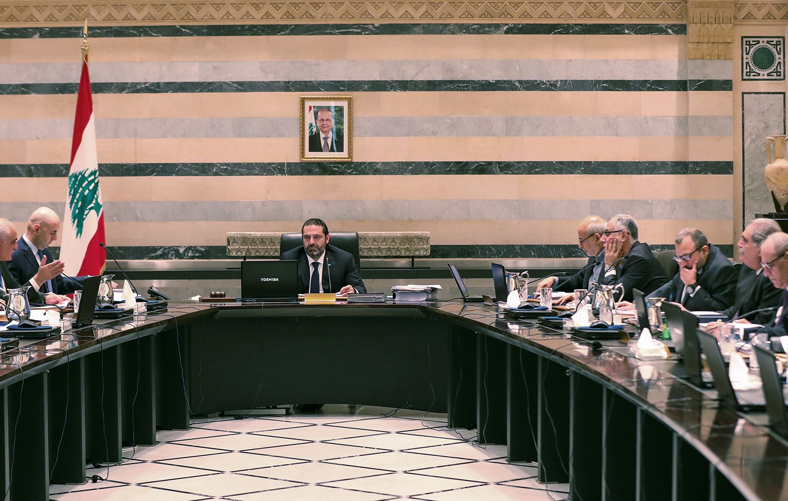 تصویر یک جلسه رسمی دولتی در لبنان را نشان میدهد. تعدادی از مقامات و اعضای دولت در اطراف یک میز بزرگ نشستهاند. در مرکز جلسه، یک مرد با لباسی رسمی و آرامش بر روی صندلی ریاست نشسته است و به دیگران نگاه میکند. در پسزمینه، پرچم لبنان و تصویری از یک شخصیت مهم بر دیوار نصب شده است. فضای اتاق با خطوط عمودی و افقی تزئینی و رنگهای ملایم طراحی شده است. افراد حاضر در جلسه به موضوعات مختلفی مشغول بحث و تبادل نظر هستند.