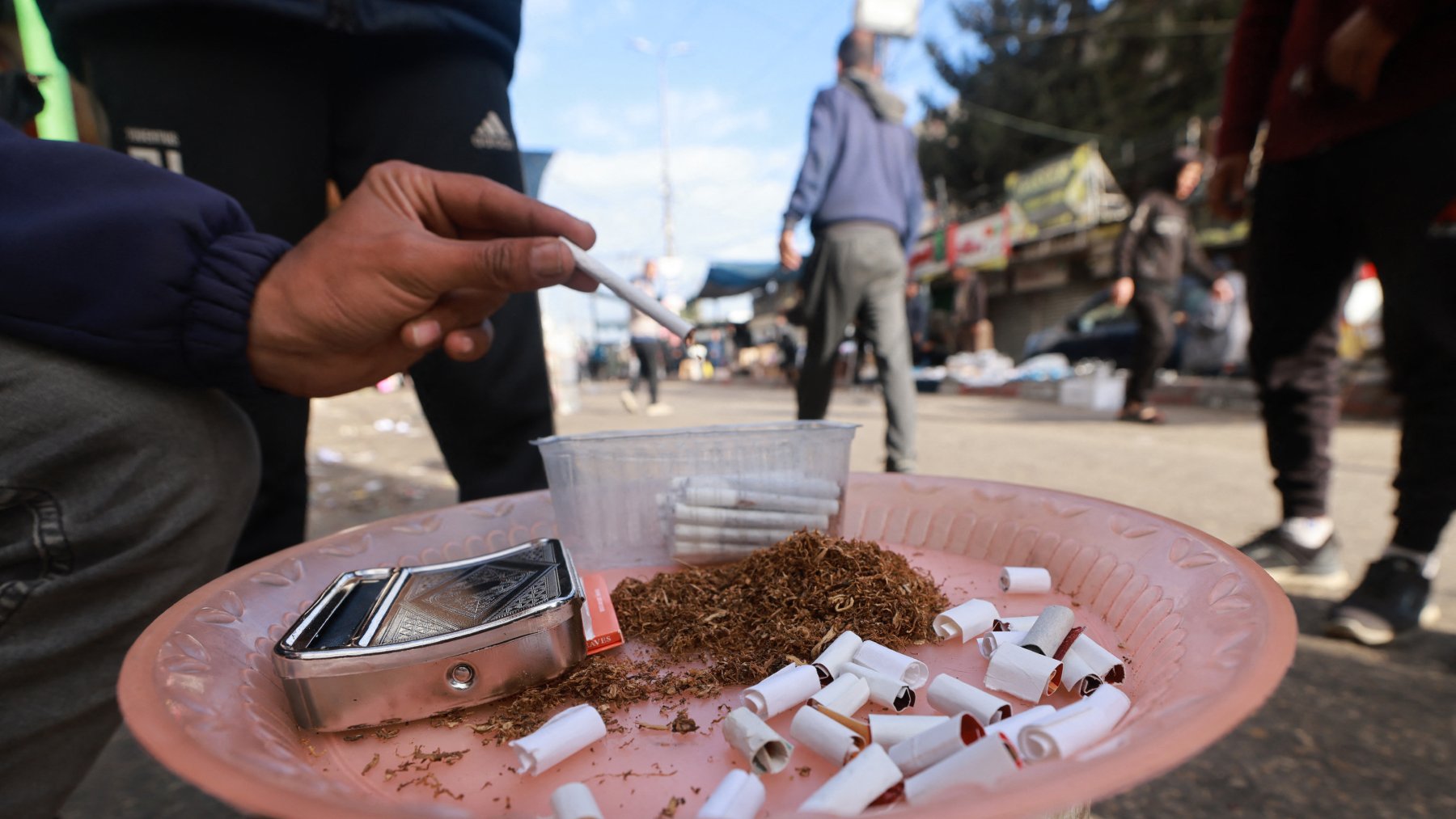 در این تصویر، یک دست در حال نشانهگذاری به سمت یک ظرف است که شامل تنباکو و سیگارهای نیمهکاره است. ظرف به صورت دایرهای و روی یک سطح قرار دارد. در اطراف، افرادی دیده میشوند که در حال عبور هستند. به نظر میرسد که این مکان یک نقطه تجمع یا خیابانی شلوغ باشد. سیگارها بههمراه تنباکو نشاندهنده فعالیت سیگار کشیدن در این معبر است.