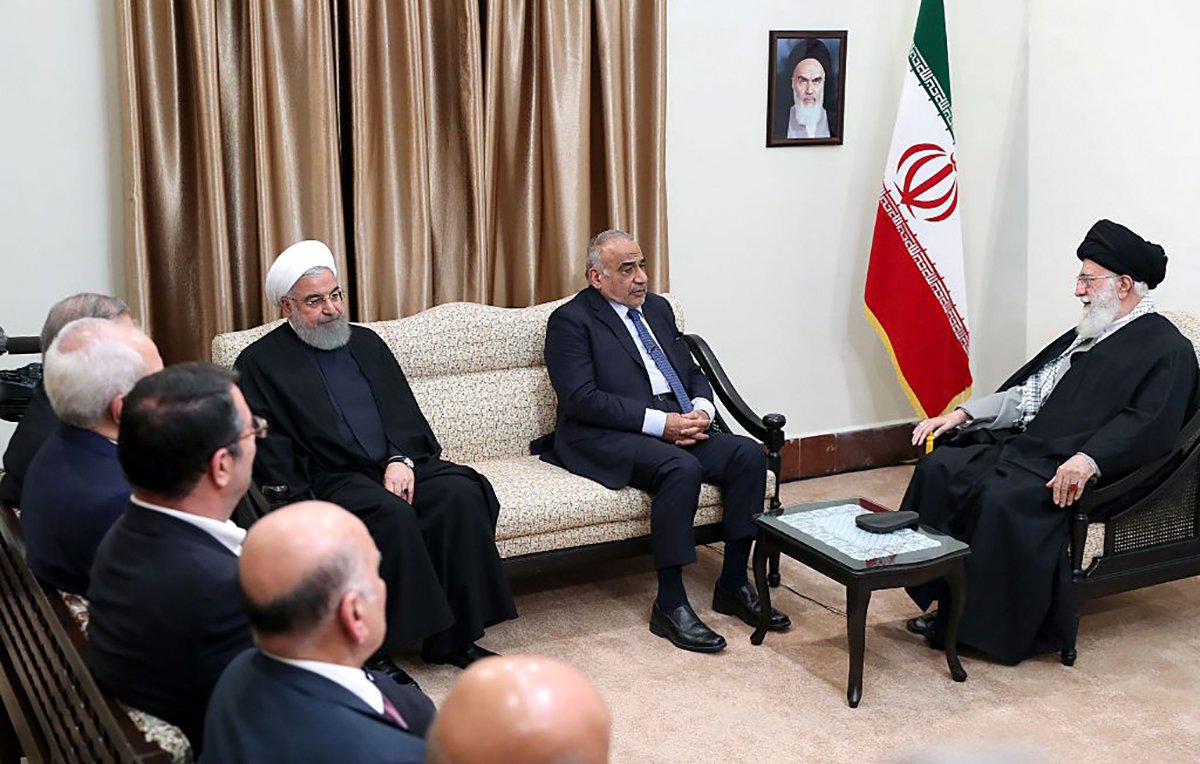 در این تصویر، چند نفر در یک اتاق رسمی نشستهاند. شخصی در سمت راست تصویر با لباس مشکی و عمامه، رهبر جمهوری اسلامی ایران است. در مجاورت او، فرد دیگری نشسته که به نظر میرسد مرتبط با مقامات دولتی باشد. در سمت چپ، افراد دیگری در حال گوش دادن به صحبتها هستند. دیوارها با پردههای قهوهای تزیین شده و پرچم جمهوری اسلامی ایران در پسزمینه دیده میشود. یک تصویر از بنیانگذار جمهوری اسلامی نیز بر دیوار نصب شده است. جو تصویر رسمی و جدی به نظر میرسد.