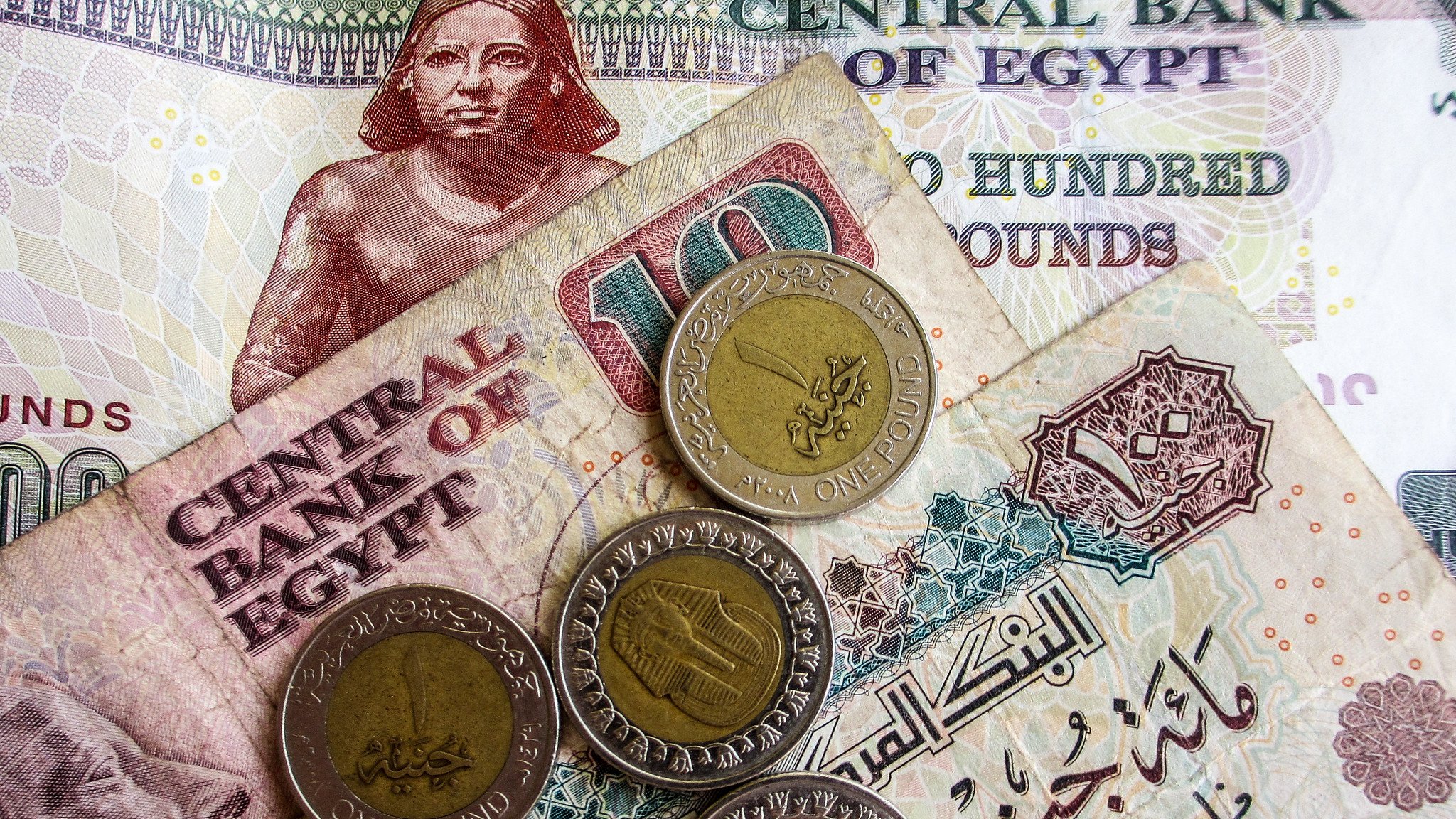 در این تصویر، تعدادی پول نقد مصری به نمایش گذاشته شده است. شامل یک اسکناس ۲۰۰ پوندی و یک اسکناس ۱۰ پوندی که در پسزمینه دیده میشود. همچنین چهار سکه یک پوندی روی اسکناسها قرار دارد. طراحی اسکناسها و سکهها حاوی نقوش و نوشتههایی به زبان عربی است که بیانگر هویت بانک مرکزی مصر و ارزش پولها میباشد. رنگهای مختلف اسکناسها و سکهها به وضوح قابل مشاهده است.