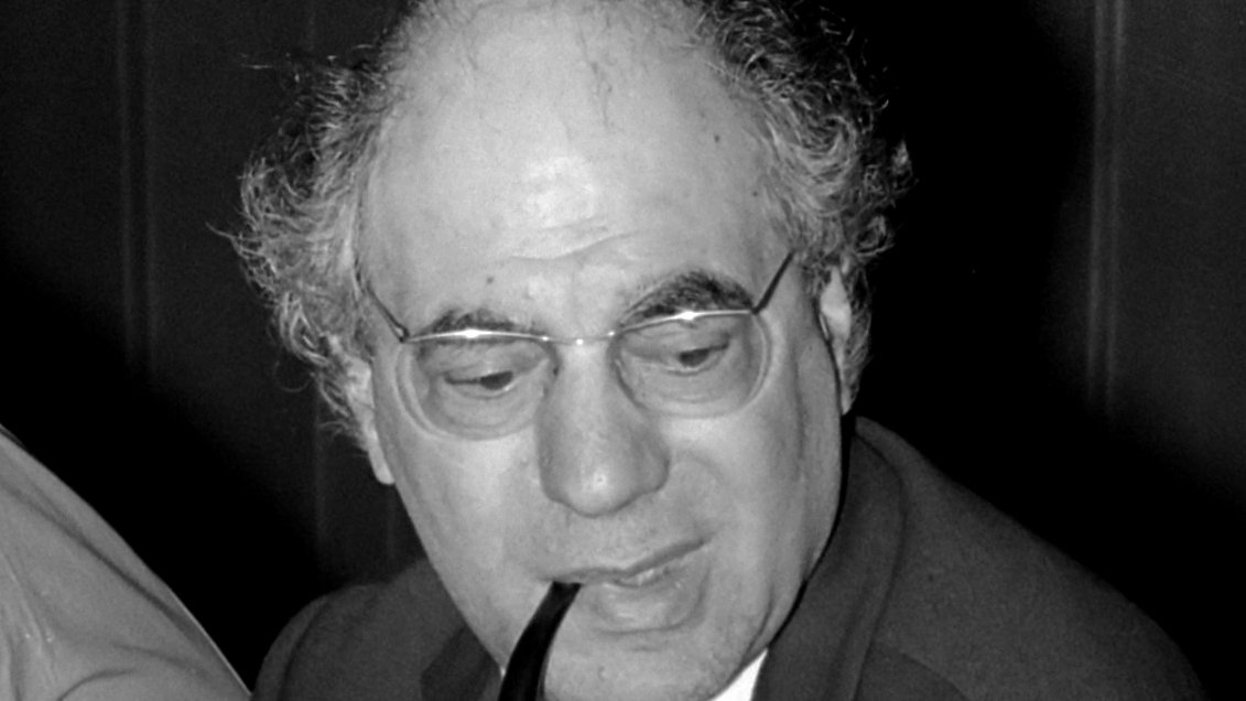 در این تصویر، فردی با موهای سفید و حالت نیمهخنثی دیده میشود. او عینکی بر چشمانش دارد و یک پیپ در دهانش قرار داده است. لباس او شامل یک کت تیره و یک کراوات است. به نظر میرسد که او در حال گفتگو یا تفکر است و توجهش به سمت دیگری متمرکز شده است. زمینه تصویر نیز تیره به نظر میرسد.