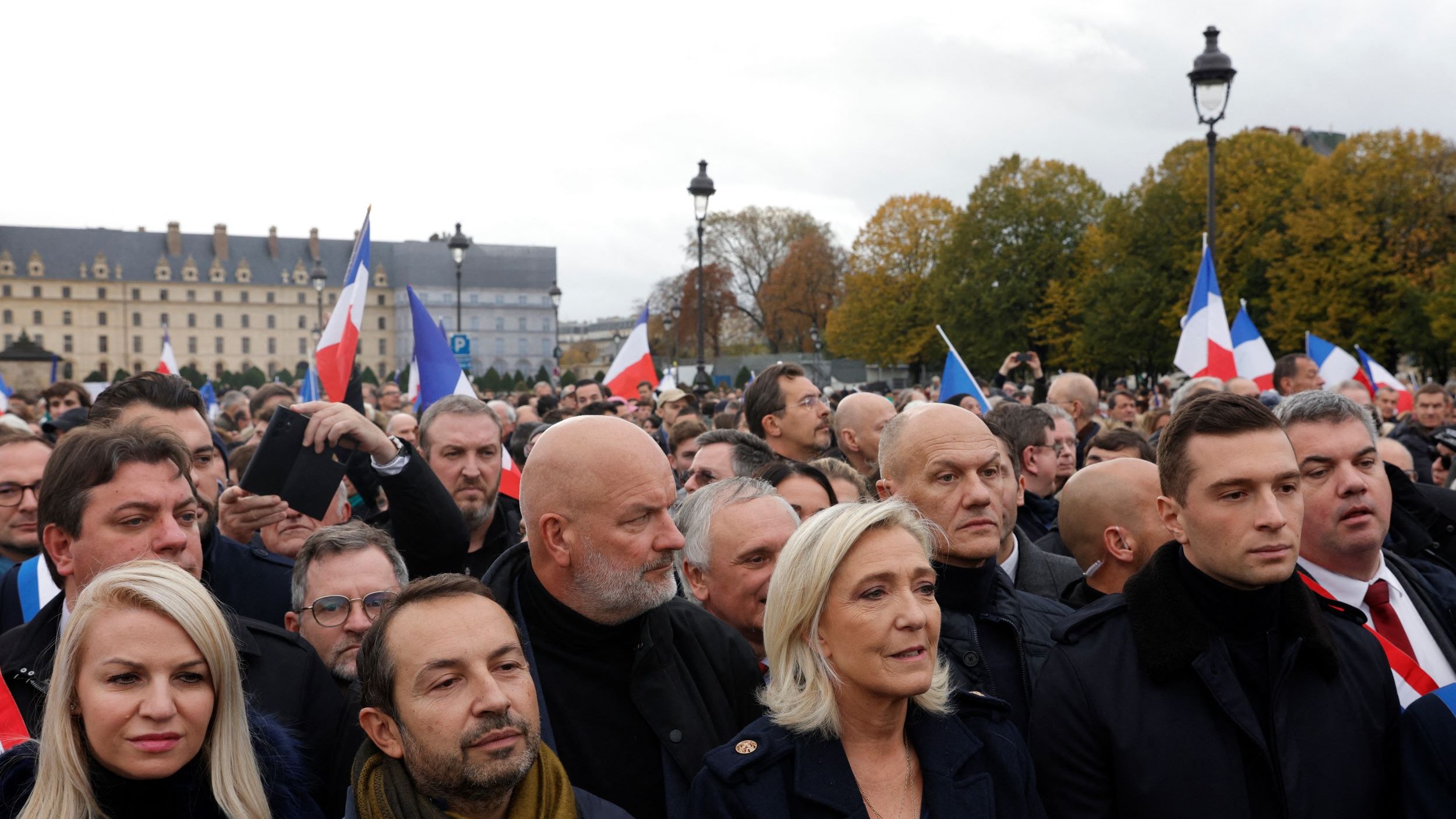 در این تصویر، گروهی از مردم در یک تجمع یا راهپیمایی در فضای باز به نمایش درآمدهاند. بسیاری از افراد پرچمهای فرانسه را در دست دارند. در میان جمعیت، افرادی با چهرههای مختلف دیده میشوند که نشاندهنده نظرات و احساسات مختلفی هستند. برخی از افراد لباسهای رسمی و گرم پوشیدهاند و به نظر میرسد موضوع این تجمع سیاسی باشد. آسمان ابری است و فضای عمومی کمی شلوغ و پرتحرک به نظر میرسد.