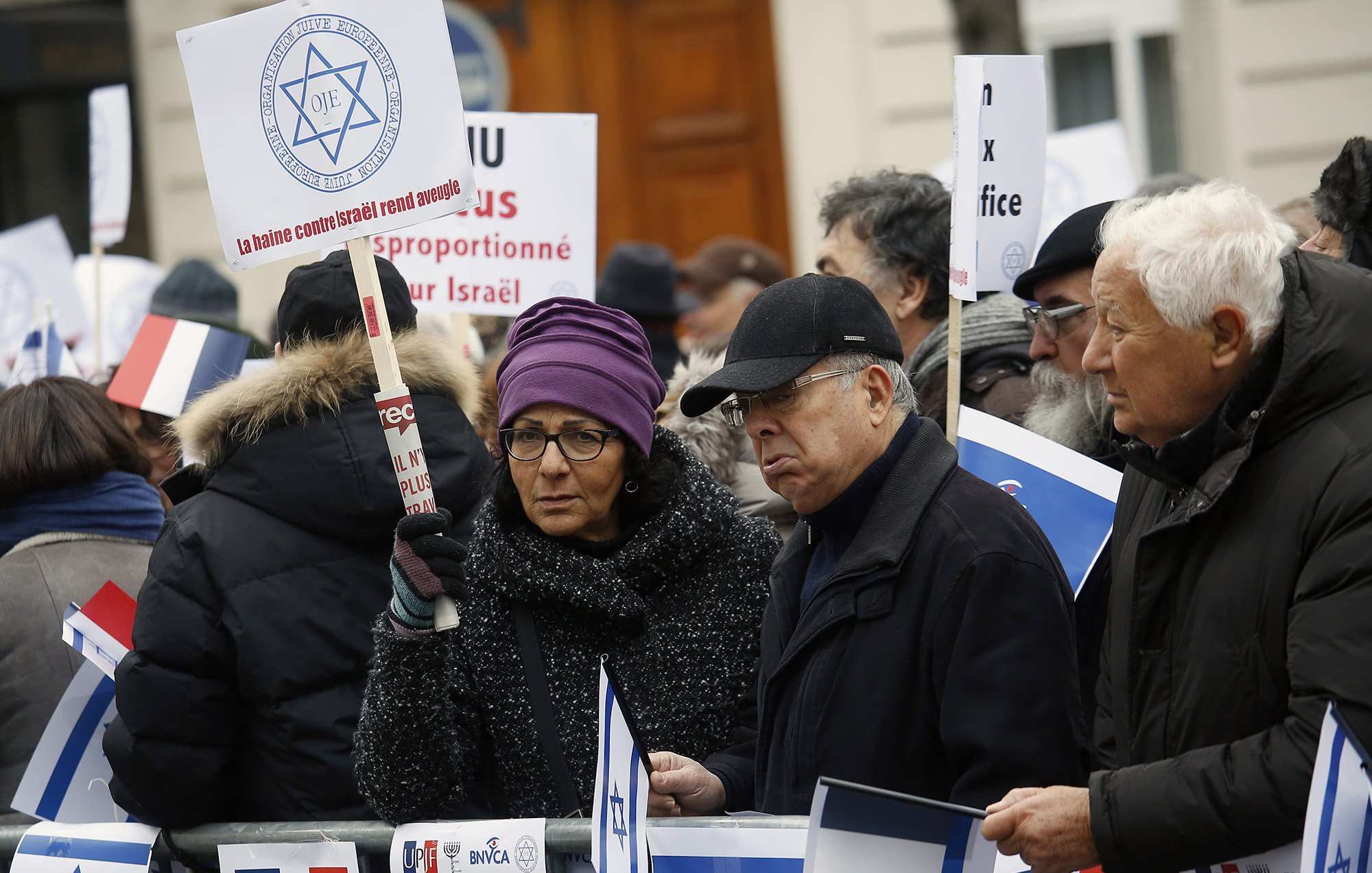 در این تصویر، گروهی از افراد در حال اعتراض هستند. آنها پلاکاردها و تابلوهای مختلفی در دست دارند. برخی از افراد لباسهای گرم به تن دارند و در میان آنها یک زن با عینک دیده میشود. تابلوها شامل پیامهایی مرتبط با اسرائیل هستند و برخی از افراد به سمت دوربین نگاه میکنند. فضای تجمع در یک مکان عمومی به نظر میرسد.