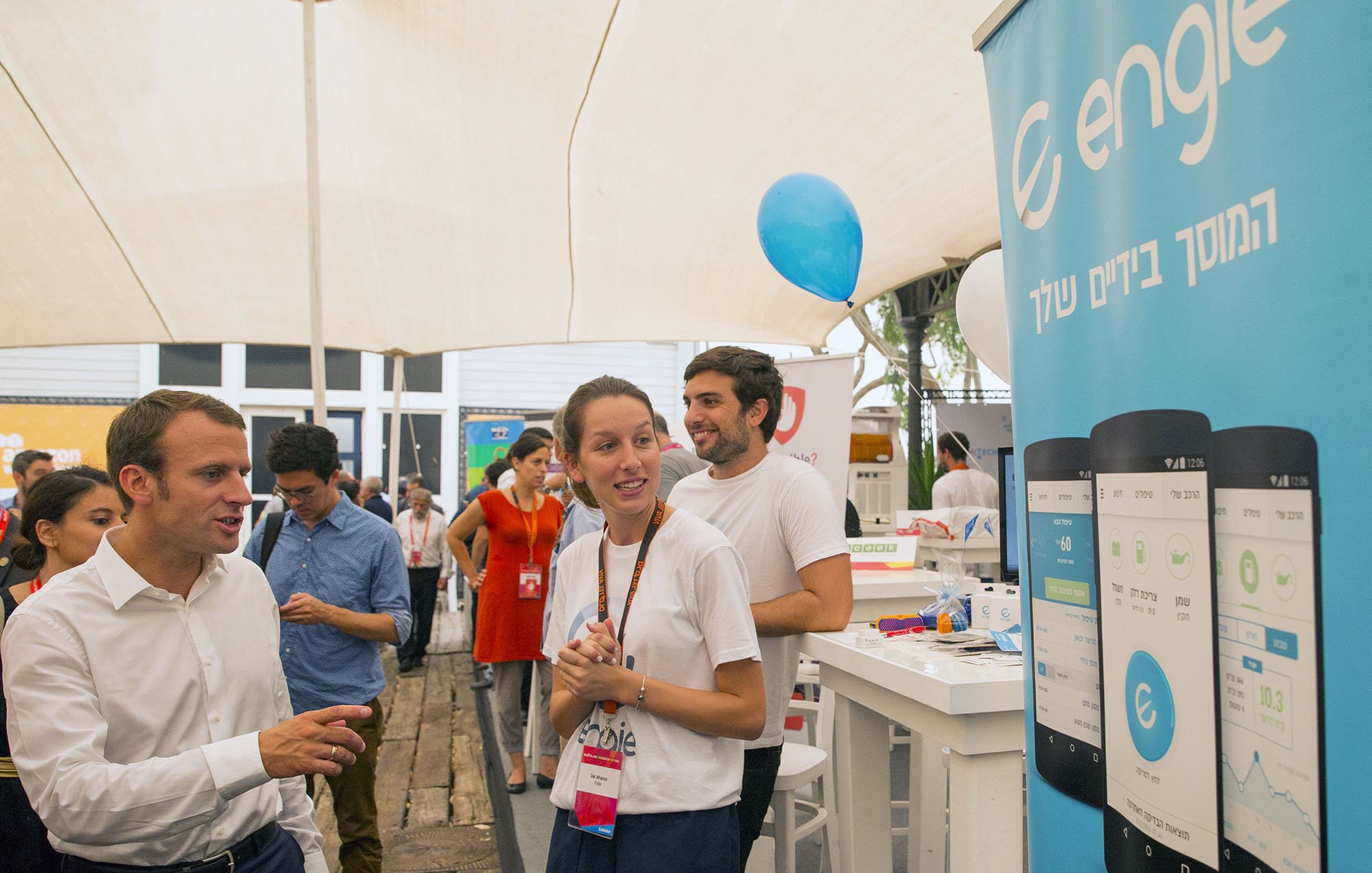 در این تصویر، یک فضای تجاری یا نمایشگاهی دیده میشود. در سمت چپ، فردی با لباس سفید و شلوار تیره در حال گفتگو است که ممکن است یک رهبر یا شخصیت مهم باشد. در سمت راست، یک زن جوان با تیشرت سفید و آرم شرکت در حال صحبت و لبخند زدن است. همچنین، در پسزمینه، افرادی دیگر در حال فعالیت هستند و یک بنر با لوگوی "engie" و تصاویری از یک برنامه موبایل وجود دارد. فضای عمومی و دوستانه به نظر میرسد.