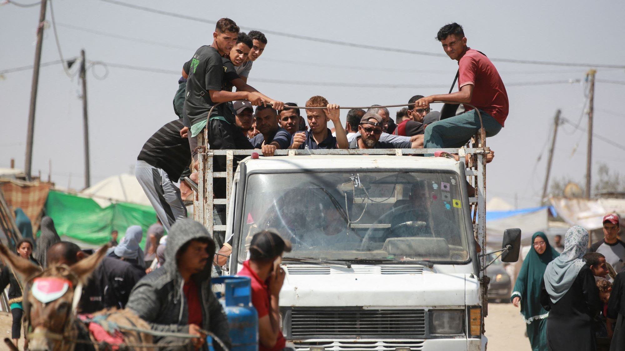 در این تصویر، گروهی از جوانان در حال نشستن بر روی یک کامیون هستند. برخی از آنها در قسمت بار کامیون نشستهاند، در حالی که دیگران به طرف خودرویی که در حرکت است، نگاه میکنند. در جلوی تصویر، چند نفر در کنار کامیون و همچنین زنانی با پوششهای سنتی مشاهده میشوند. در پسزمینه، سایههایی از چادرها و همچنین سیمکشیهای برق دیده میشود که نشاندهنده یک منطقه پرجمعیت و شاید غیررسمی است. احساسی از شلوغی و جنب و جوش از صحنه بهطور کلی مشهود است.