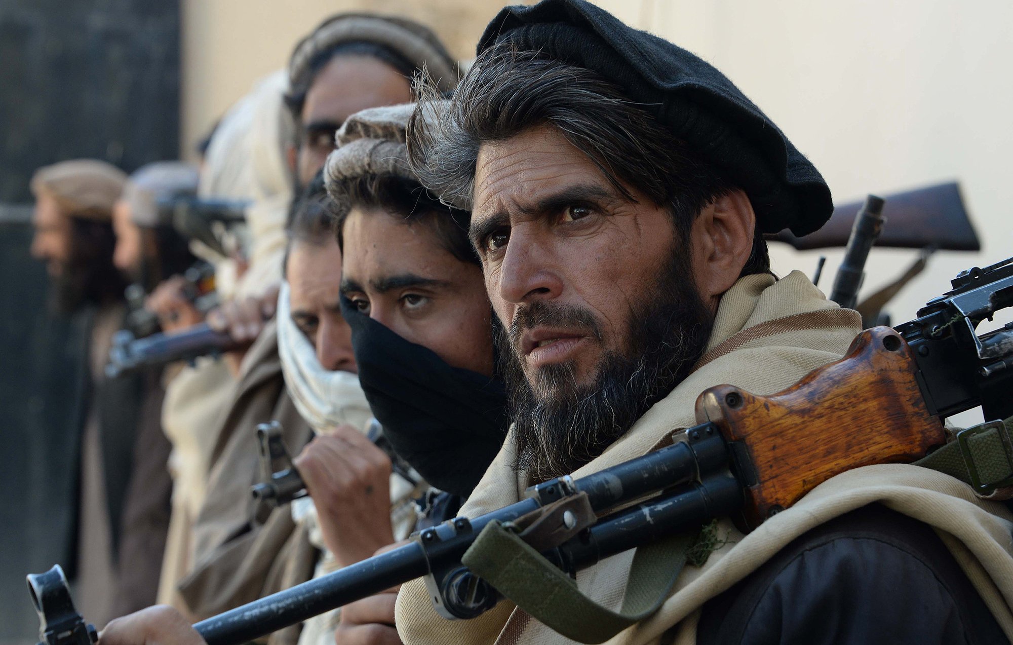 این تصویر گروهی از مردان مسلح را نشان میدهد که در یک وضعیت آمادهباش قرار دارند. آنها لباسهای سنتی دارند و بعضی از آنها صورت خود را با پارچه پوشاندهاند. سلاحهایشان بر دوششان قرار دارد و تمرکز و جدیت آنها در چهرهشان مشهود است. زمینه فقط به صورت محو دیده میشود و توجه بیشتر به افراد معطوف است.