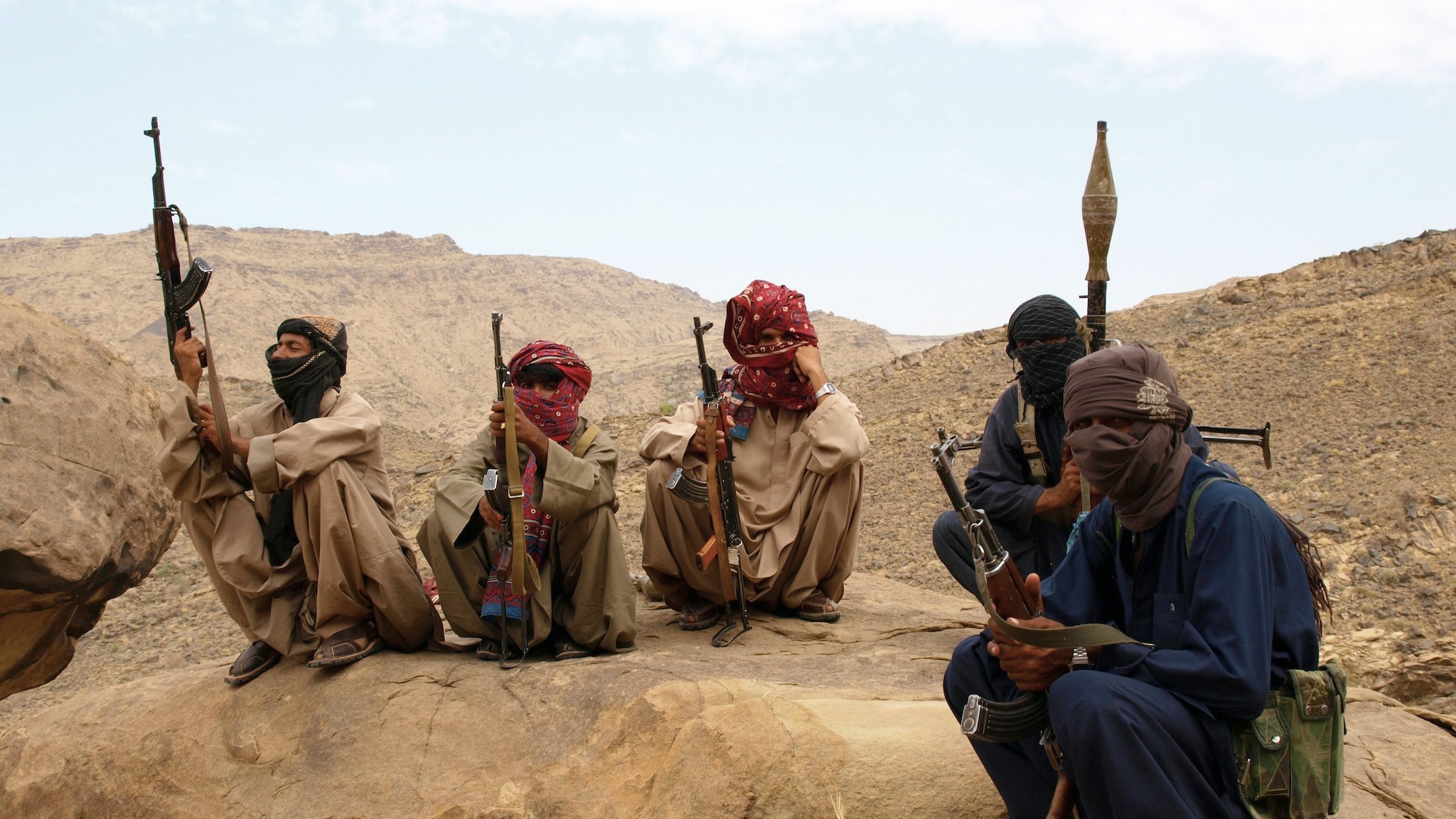 تصویر گروهی از مردان مسلح را نمایش میدهد که در یک منطقه بیابانی یا کوهستانی نشستهاند. آنها لباسهای سنتی محلی به تن دارند و صورتهایشان با پوشاکی مثل شال یا دستمال پوشانده شده است. برخی از آنها تفنگها و سلاحهای دیگری در دست دارند، در حالی که در حال نظارت بر محیط اطراف خود هستند. پسزمینه شامل سنگها و تپههای بیابانی است. وضعیت عمومی تصویر حس جدیت و آمادگی را منتقل میکند.