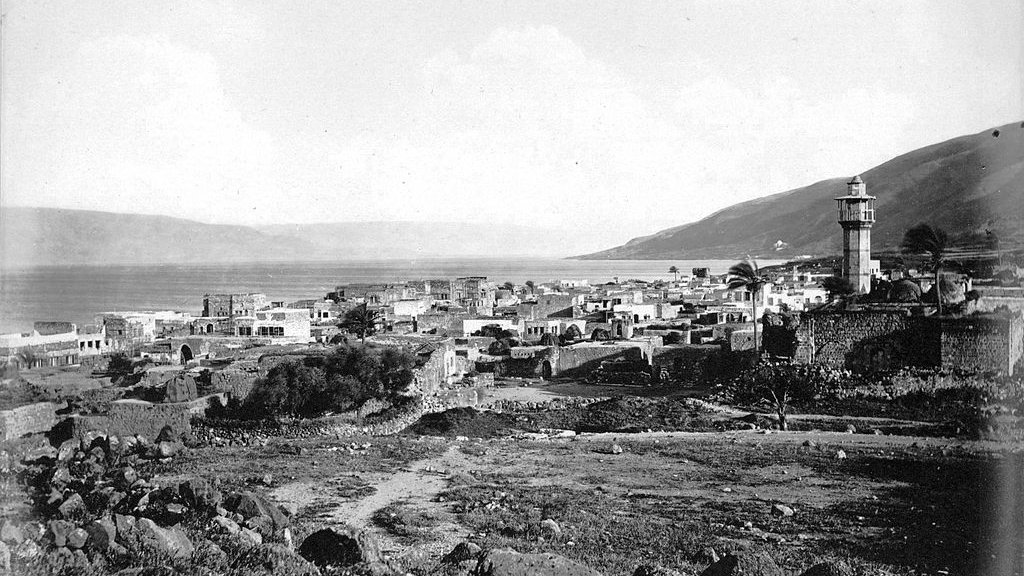 این تصویر نمایشدهنده یک مناظر تاریخی از یک روستا یا شهر نزدیک به دریاچه است. در تصویر، یک برج باستانی در سمت راست دیده میشود که به واسطه آن، هویت محلی خاصی به چشم میآید. ساختمانها در سمت چپ و در پسزمینه قرار دارند و برخی از آنها به نظر میرسد که با مصالح سنگی ساخته شدهاند. در دوردست، کوهها و زمینهای مرتفع قابل مشاهدهاند و ابرها در آسمان پراکندهاند. به طور کلی، این تصویر حس تاریخ و طبیعت آرام را به بیننده منتقل میکند.