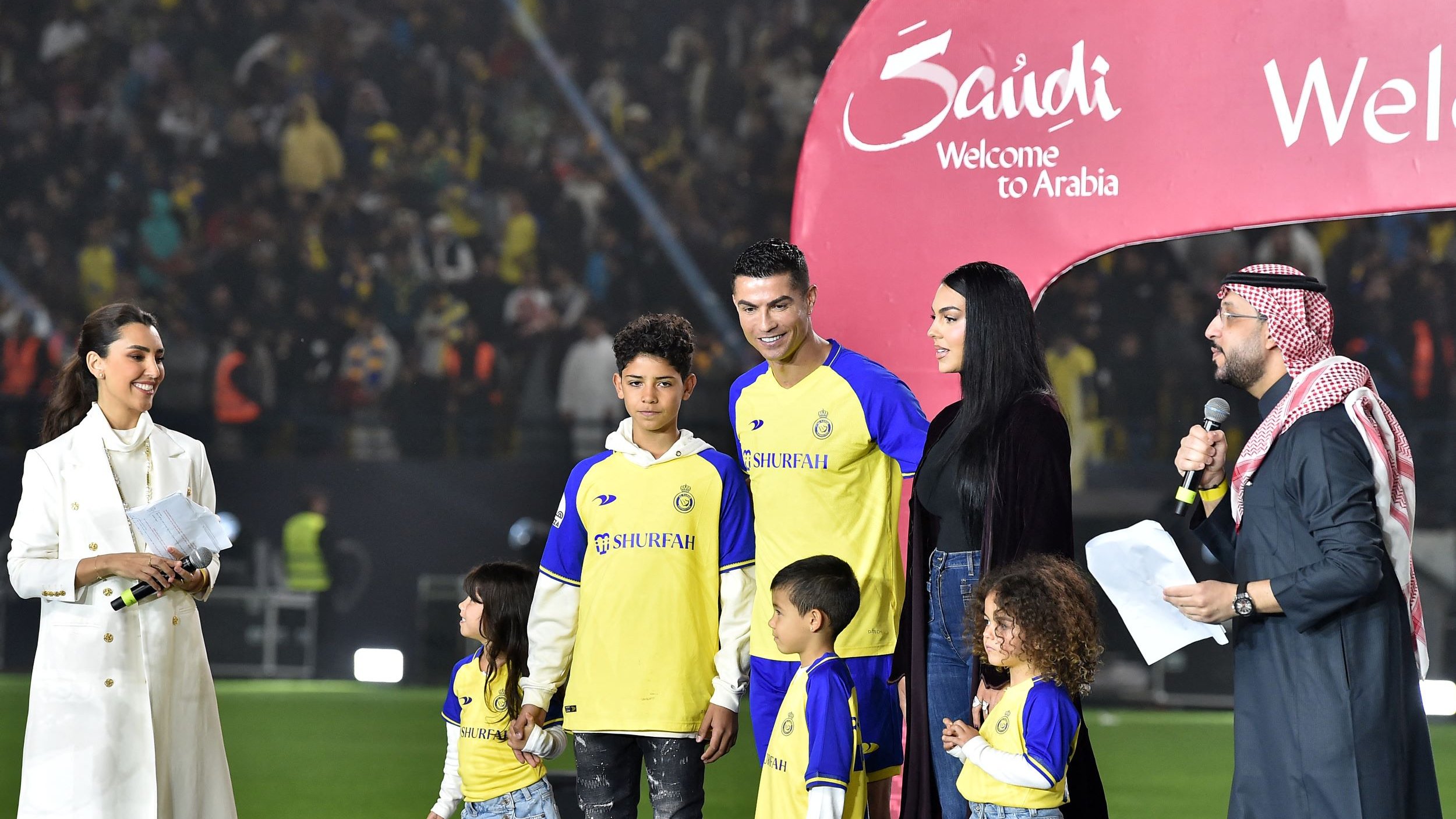 در این تصویر، یک رویداد ورزشی در حال برگزاری است. در وسط تصویر، کریستیانو رونالدو، بازیکن معروف فوتبال، همراه با چند کودک و یک زن ایستاده است. آنها لباسهای تیمی با رنگ زرد و آبی پوشیدهاند. در پسزمینه، جمعیتی از طرفداران به وضوح دیده میشوند. همچنین، یک نفر در حال صحبت در میکروفون است و دو نفری که سمت چپ تصویر قرار دارند، به نظر میرسد که مجری برنامه هستند. به نظر میرسد که این رویداد به معرفی یا خوشامدگویی به رونالدو مربوط میشود. یک بنر بزرگ با نوشته "Welcome to Arabia" نیز در تصویر دیده میشود.