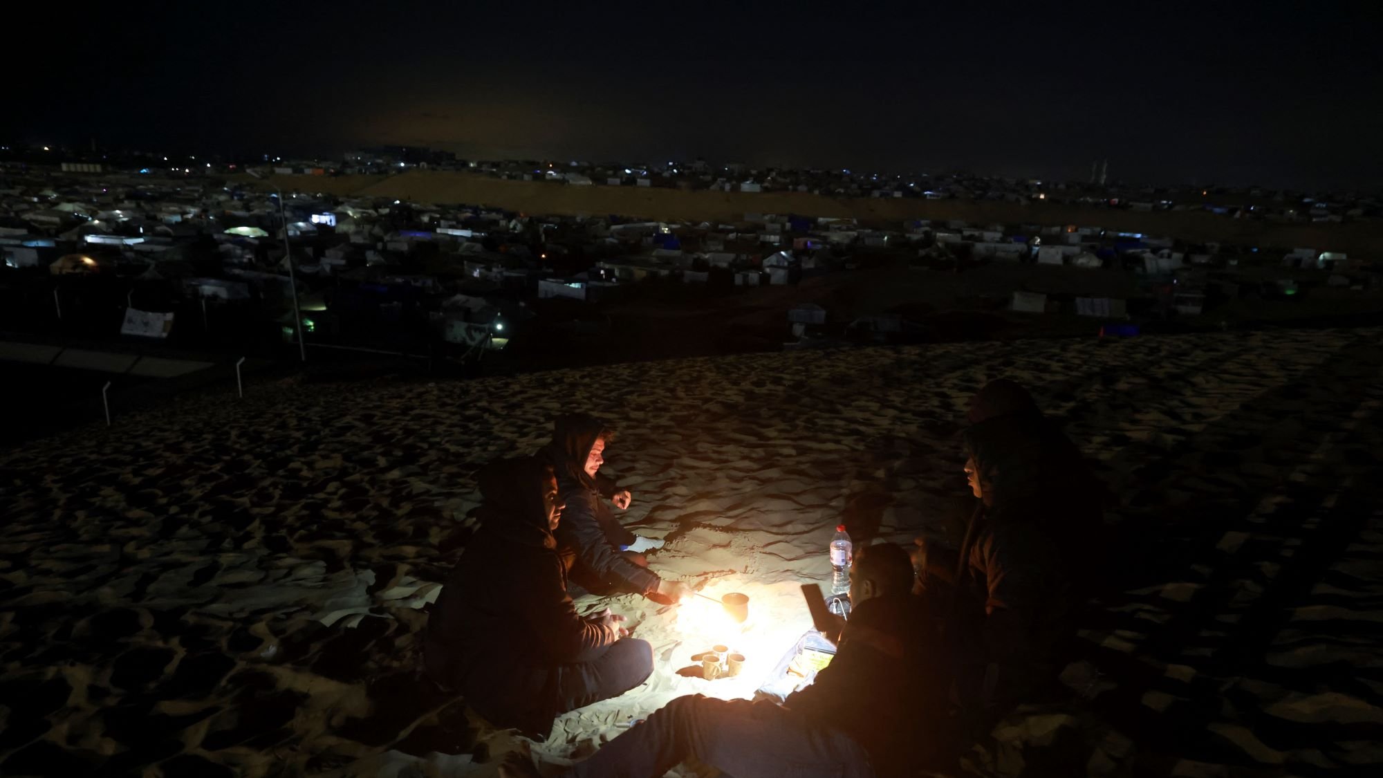 در این تصویر، یک گروه از افراد در شب در حال دور هم نشستن در بیابان هستند. آنها در اطراف آتش نشستهاند و نور ملایم آتش نور را بر چهرههایشان میتاباند. در پسزمینه، میتوان چادرها و نورهای دیگری را که در دوردست قرار دارند، مشاهده کرد. این صحنه حس صمیمیت و نزدیکی به طبیعت را منتقل میکند. همچنین، فعالیتهای شبانه و سکوت بیابان به فضای تصویر عمق بیشتری میبخشد.