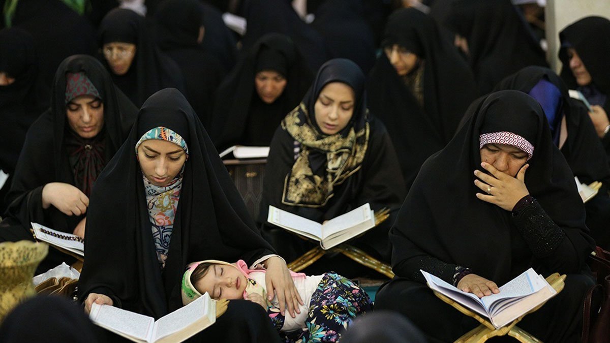 در تصویر گروهی از زنان با چادرهای سیاه در حال مطالعه قرآن دیده میشوند. بعضی از آنها در حال خواندن هستند و توجه عمیقی به کتاب دارند. همچنین، یک کودک در آغوش یکی از زنان خوابیده است. بهطور کلی، فضایی معنوی و پر از تمرکز در این تصویر وجود دارد.