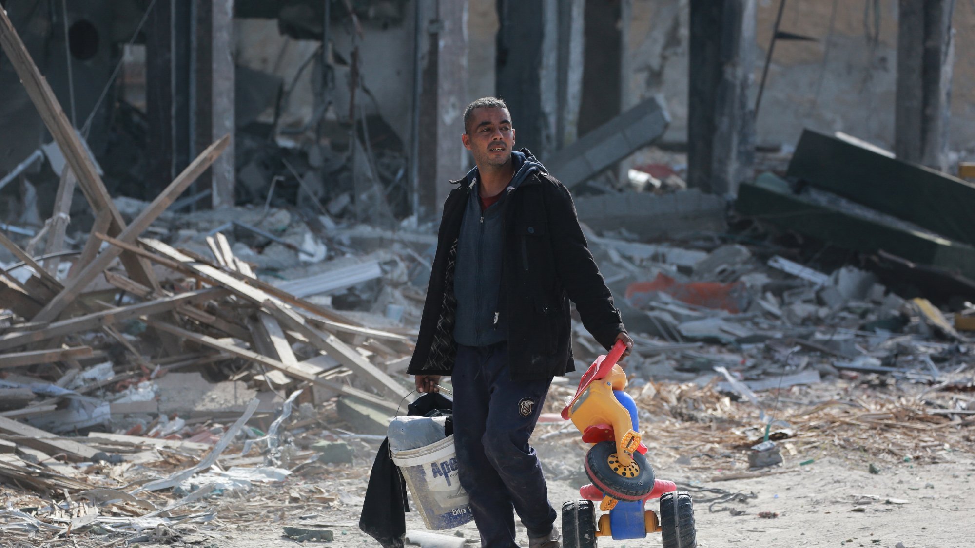 تصویر مردی را نشان میدهد که در فضایی ویران شده و پر از آوار حرکت میکند. او با ظاهری خسته، یک سطل در دست دارد و در دست دیگرش یک اسباببازی رنگی که شبیه به دوچرخه است، حمل میکند. پسزمینه تصویر شامل بقایای ساختمانها و مواد ساختمانی پراکنده است که نمایانگر وضعیت ناراحتکنندهای است.