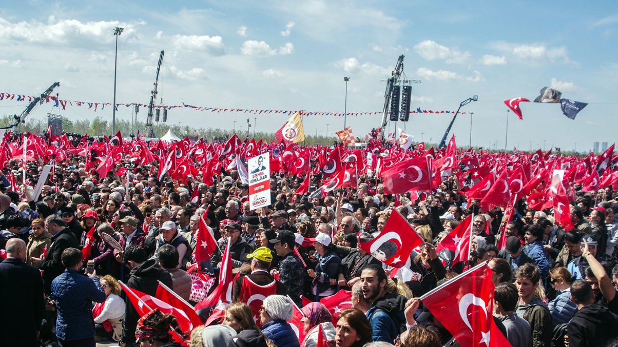 تصویر نشاندهنده تظاهرات بزرگی در یک فضای باز است که تعداد زیادی از مردم حضور دارند. آنها پرچمهای قرمز و سفید ترکیه را در دست دارند و به نظر میرسد که در حال ابراز همبستگی یا حمایت از یک حرکت یا رویداد خاص هستند. آسمان آبی است و چند ابر سفید در آن دیده میشود. جمعیت متنوعی در تصویر وجود دارد که نشاندهنده حضور افراد با سنین و زمینههای مختلف اجتماعی است. فعالیت و شور و حال در این جمعیت به وضوح قابل مشاهده است.
