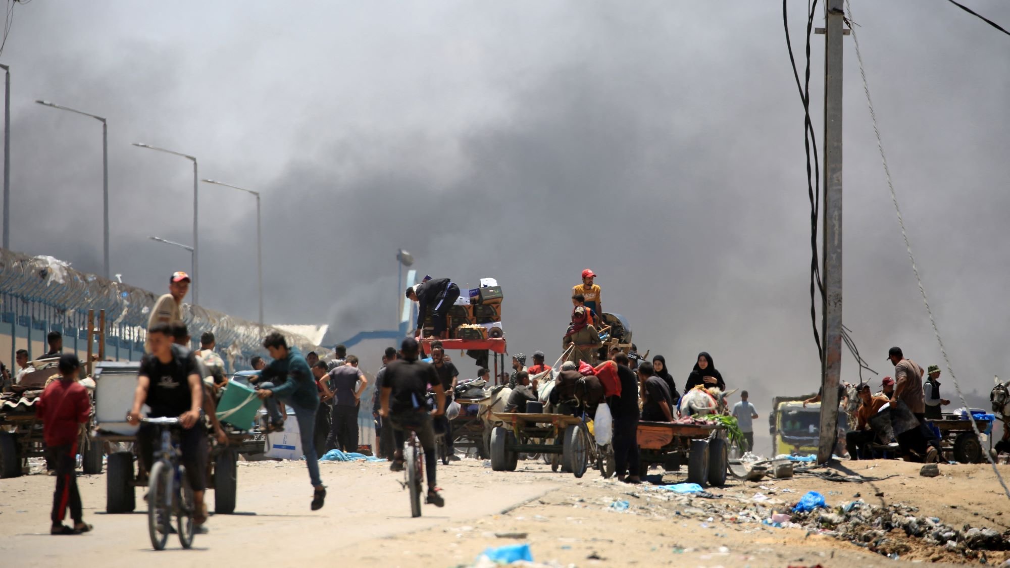 تصویر یک صحنه شهری را نشان میدهد که در آن تعدادی از افراد در حال حرکت هستند. در زمینه، دود سیاهی به آسمان بلند شده که نشاندهنده وجود یک حادثه یا آتشسوزی است. افرادی که در تصویر دیده میشوند، شامل مردان، زنان و کودکانی هستند که برخی از آنها در حال دوچرخهسواری و برخی دیگر در حال حمل بار در کالسکههای دستی هستند. فضای کلی تصویر به نظر شلوغ و پر از فعالیت است.