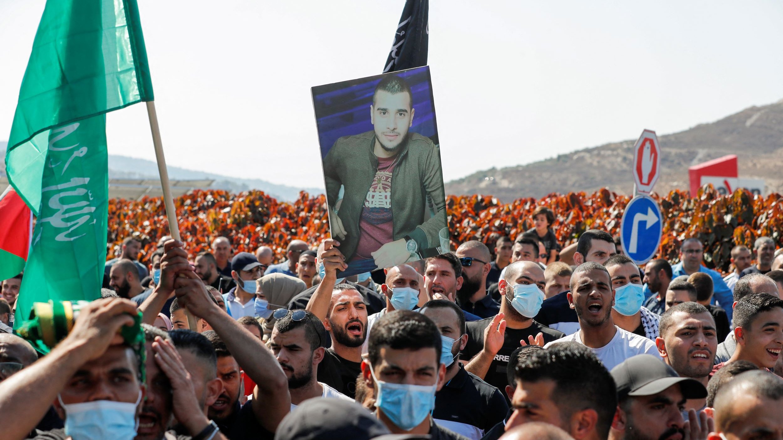 در تصویر، گروهی از افراد در یک تظاهرات حضور دارند. تعداد زیادی از افراد پرچمهایی به رنگهای مختلف در دست دارند. یکی از تظاهرکنندگان تصویری از یک مرد را بالا نگه داشته است. بیشتر افراد ماسک به صورت دارند و در پسزمینه، زمینهای سرسبز و تپههایی دیده میشود. به نظر میرسد که این تجمع به منظور اعتراض یا یادبود برگزار شده باشد.