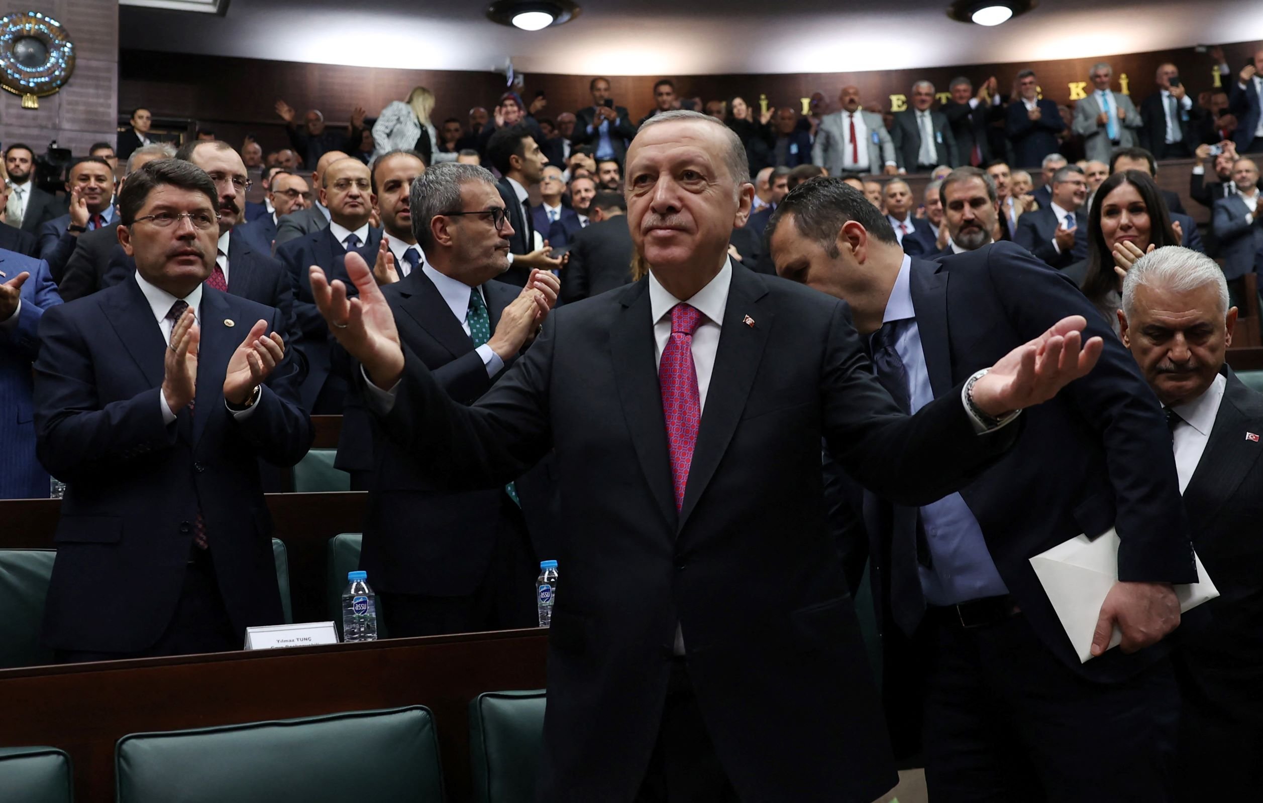 در این تصویر، یک جمعیت بزرگ از افرادی که در یک سالن جلسه نشستهاند، دیده میشود. در مرکز تصویر، فردی با لباس رسمی و کراوات صورتی در حال خوشآمدگویی یا ابراز احساسات است. او با دستان باز به سمت مخاطبان اشاره میکند. در پسزمینه، اعضای دیگر نیز ایستاده و به او نگاه میکنند. برخی از آنها در حال تشویق و applauding هستند. فضای کلی مراسم جدی و رسمی به نظر میرسد.