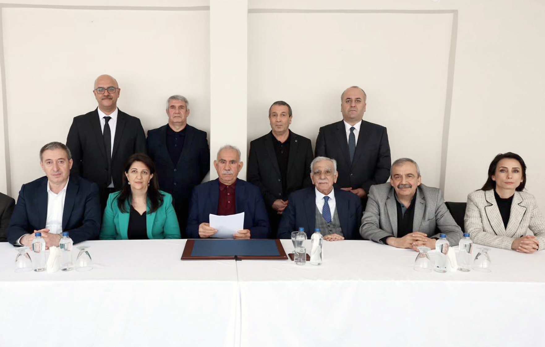 تصویر یک گروه از افراد را نشان میدهد که در یک جلسه رسمی نشستهاند. آنها به ترتیب در دو ردیف قرار دارند. میز روبروی آنها خالی از اشیا است و در پسزمینه، دیواری ساده و روشن وجود دارد. افراد لباسهای رسمی پوشیده و به دوربین نگاه میکنند.