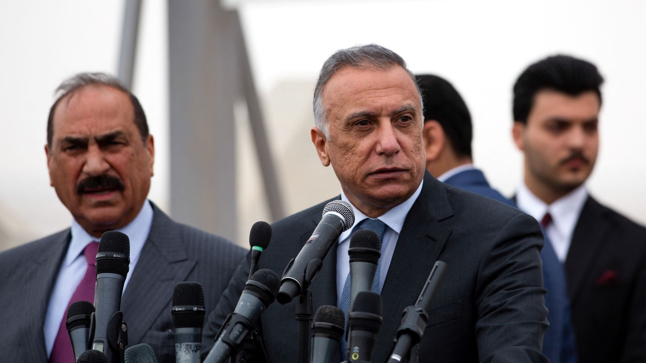 در این تصویر، دو فرد در یک کنفرانس خبری یا مراسم رسمی دیده میشوند. فردی که در سمت راست قرار دارد، لباس رسمی پوشیده و به نظر میرسد در حال پاسخ دادن به سوالات یا صحبت کردن است. او چند میکروفن مقابل خود دارد که نشان از اهمیت موضوع دارد. فرد دیگر در سمت چپ نیز لباس رسمی دارد و در حال گوش دادن به صحبتهای فرد اول است. پسزمینه تصویر کمی مبهم به نظر میرسد. وضوح و جزئیات چهرهها به دلیل زاویه و ترکیب تصویر محدود است.