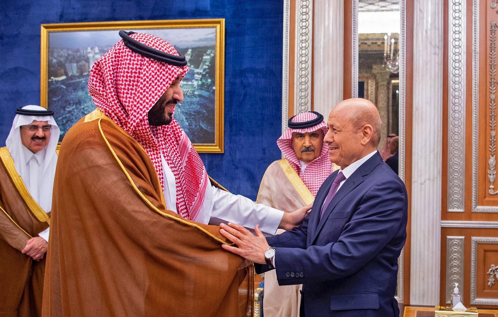 در این تصویر، دو مرد در حال گفتوگو و خوشآمدگویی به یکدیگر هستند. یکی از آنها لباس سنتی عربی به نام "ثوب" به تن دارد و چفیهای خاص بر سر دارد. او به نظر میرسد که در مقام رهبری قرار دارد. مرد دیگر نیز به صورت رسمی لباس پوشیده است و نشان از احترام و ادب در بیان حرکات او دیده میشود. در پسزمینه، دکوراسیون مجلل و لوکس با تزئینات طلایی و تابلوهایی از مناظر شهری دیده میشود که به فضای رسمی این ملاقات افزوده است.